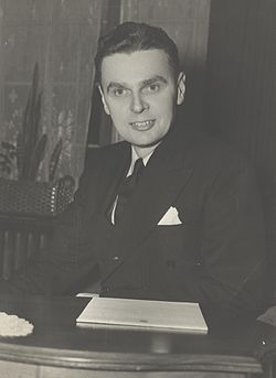 Diefenbaker, with a pleased expression on his face, sits at a desk, before him is a pile of House of Commons stationary.