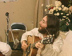 A bearded, bespectacled man in his late twenties, with long black hair and wearing a loose-fitting pajama shirt, sings and plays an acoustic guitar. White flowers are visible behind and to the right of him.