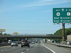 A mulitlane freeway in an urbanized area with two sets of roads in each direction. A green sign is visible in between the lanes on the right side of the road reading exit 9 2 miles Route 18 U.S. Route 1 New Brunswick East Brunswick