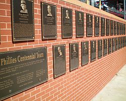 A succession of black metal plaques mounted on a brick wall. In the foreground is one plaque larger than the other entitled "Phillies Centennial Team". The smaller plaques each have a face and inscribed text.