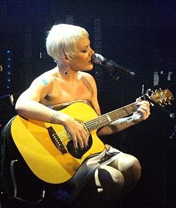 A woman with short blonde hair sitting on a chair, playing guitar and singing into a micophone