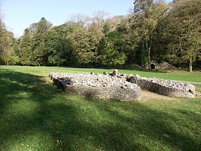 Front view of a cairn, from its right side, its boulders retained by a short wall that forms a courtyard at its entrance. The cromlech is set in flat ground of short grass (in dappled sunlight in the foreground and full sun elsewhere), dissected by a path passing behind it. Trees are mainly in leaf to its rear, among which a limestone kiln is visible at the foot of a gorge.