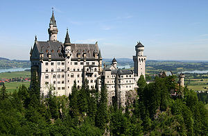A castle of fairy-tale appearance sitting high on a ridge above a wooded landscape. The walls are of pale stone, the roofs are of steep pitch and there are a number of small towers and turrets.