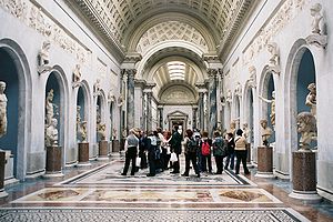 Photo of a gallery in the museum. It is in the Classical style and has a wide arched roof with skylights. The colour scheme is are blue-grey and white with a polychrome marble floor. The walls of each side of the gallery have a row of large niches in which stand marble statues. Between the niches are plinths supporting smaller portrait sculptures.