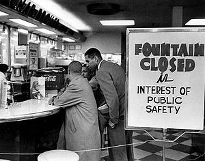 Lunch counter with four young black men seated at bar stools along the counter. They are not being served and are simply sitting. A sign on the checkerboard-tiled floor reads "Fountain closed in interest of public safety".
