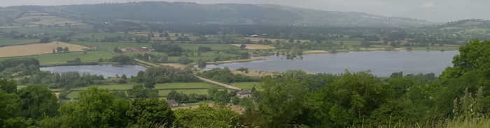 photograph of the lake showing a road crossing via a causeway at Herriots Bridge. It is surrounded by green vegetation with hills in the distance
