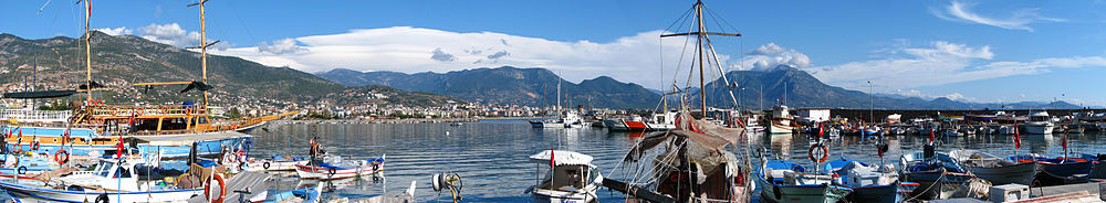 Numerous white, blue, and orange boats float tied to a dock in a greenish sea. Mountains rise on the other side of a bay.