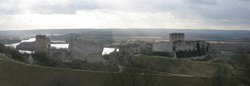 The ruins of a castle in grey limestone dominating the landscape. The River Seine is in the background. The castle's keep protrudes above the walls of the inner bailey on the right, with a bridge leading up to the bailey's entrance. To are ruins of the wall enclosing the outer bailey; a tower stands taller than the ruined walls.