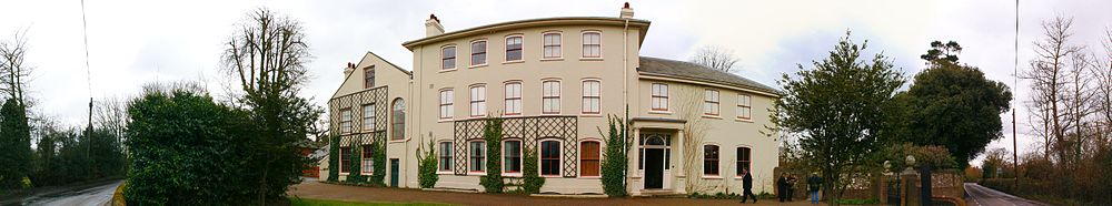 Panorama of a three storey stucco rendered building in a plain Georgian style, with bushes and trees on each side where a boundary wall shelters the gravelled garden from a narrow lane.