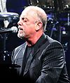 A man with a goatee and his eyes closed behind a microphone on a piano, wearing a black shirt and striped jacket.