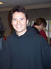 A man in his early 30s wearing a plain, dark colored hooded sweatshirt, smiling