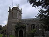 Stone building with square tower at left hand end. In the foreground are trees and gravestones.