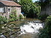 Water flowing through a channel and over a weir between a building and a wall. Vegetation on both sides of the water.