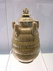 A tan jar with several rounded bands carved into the body and five upward facing tubes equadistantly spaced along the top area of the jar. The lid is crowned with a lotus flower carving.