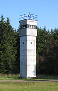 A square greyish tower on a meadow, with trees in the background. Only the topmost section has windows; there is a searchlight on the tower's flat roof, and a railing around the roof's outer edge.