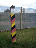 A square wooden post with diagonal black, red and yellow stripes and a metal sign saying "DEUTSCHE DEMOKRATISCHE REPUBLIK" stands in front of a steel mesh fence, through which a guard tower is visible.