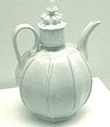 A white teapot with an almost perfectly spherical body and a large, cylindrical cap in the center which is topped with a small crown shaped embellishment. Several vertical lines are glazed into the body of the pot.