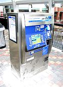 A silver and blue ticket vending machine on a station platform with payment options and digital screen visible.