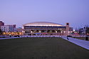 The glass-clad and stone-faced exterior of an arena in the background, with a large expansive open grass park in the foreground.