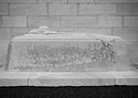 A sword, army helmet and laurel sit on top of a stone sarcophagus that is located in front of a stone wall.