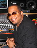 An Afro-American man is smiling. He is sitting in a control room, which includes a mixing console and monitor speakers. He wears sunglasses, a black shirt and a wristwatch.