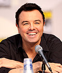 A man with black hair and a black shirt, leaning forward, smiling into a microphone