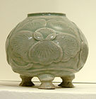 A blue-green tinted grey pot with a wide bottom supported by three stubby legs, a wide body, and a smaller opening at the top. A large five petal flower pattern is out-dented on the body of the jar.