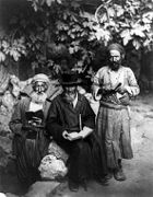 A sepia photograph shows three elderly Jewish men sporting beards and holding open books, posing for the camera. Against a backdrop of leafy vegetation, the man in the centre sits, wearing a black hat and caftan, while the two others stand, wearing lighter clothes and turbans.