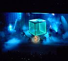 Blue lit stage with a large transparent cube in the middle, and smoke surrounding it. a number of people are dancing around the cube, wearing black dress