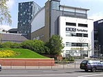 A quirky modern five-storey building with a large sign saying "BBC Yorkshire" in black above the second-floor windows on the white-fronted facade of the lower four floors can be seen on the far side of a dual-carriageway road with a barrier along the central reservation. At right-angles to the right of the building is a tall blue slab with the letters "BBC" in white at the top. The left side of the building is mostly brick-red with a few windows, but above it is a light blue windowless section. The roof above this and the grey fifth floor of the frontage curves gently down to the rear. A lone car is driving from left to right along the road;  between it and the building, temporary boards have been erected in front of a building to the left. In the top left-hand corner of the picture, part of a tall many-windowed building can be seen.