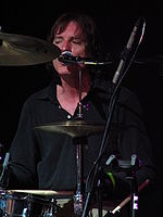 The drummer in a close-up view through the gaps between his drums and cymbals. His face and neck are slightly reddened from his exertion. While drumming, he is singing into the microphone mounted close to his face