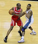 A basketball player, wearing a red jersey with the word "ROCKETS" in the front, is holding the basketball while another basketball player, wearing a white jersey, attempts to steal the ball.