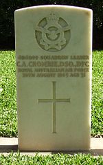 A white grave stone marker. The stone contains an insignia at the top with a crown, flying eagle and banners, followed by text of the person and a Christian cross.