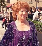 Woman in her sixties, with short, curly red hair, wearing a purple gown.