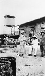 Three men in uniform, two sitting and one standing, outside a number of buildings and earthworks.
