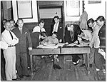 A soldier is sitting at a wooden table, with two men signing papers at the table and a line of men lined up on the right. On the left are two men, one in shirt and tie and the other in a suit facing the camera.