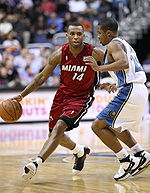 A basketball player, wearing a red jersey with the word "MIAMI" and the number 14 in the front, is dribbling the basketball while another basketball player, wearing a white jersey, attempts to defend him.