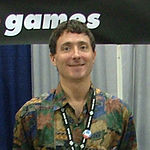 A Caucasian man with short brown hair and a convention pass on a lanyard around his neck smiles for a camera. Part of the Telltale Games logo is visible in the background.