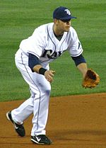 A man in a white and blue baseball uniform with the letters "TB" on his cap kneels, preparing to field a ground ball.