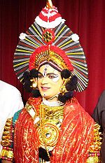 Person with painted eyes in Yakshagana costume, as gold-spangled robe with red sheer scarf and spiked headress on gold crown