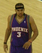 A basketball player, wearing a purple jersey with the word "PHOENIX" and the number 3 on the front, stand on a basketball court.