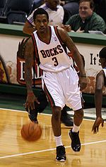 A man, wearing a green jersey with a word "BUCKS" and the number "3" written in the front, is dribbling a basketball.