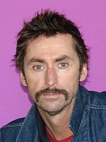 A man with brown hair and a brown mustache wearing a blue denim jacket looks directly ahead.