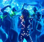 A blond woman dancing in a blue dress, which has small glowing lights on it. She is surrounded by dancers in silver, body-hugging dress with a neon green mask in front