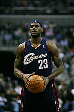 A man, wearing a black jersey with a word "CLEVELAND" and the number "23" written in the front, is holding a basketball.