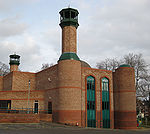 A striking modern building of two shades or red-brick with, on its left, two tall circular towers with minarets and a low entrance-building. The near end has two long thin windows with green panels and pointed tops to the right of the nearer tower. To the right of the windows is another, lower, tower with a small green dome. In the centre of the building, part of a larger dome can be seen. In the background are some trees and a red-brick Victorian terrace.