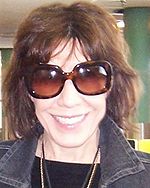 Woman in her late 1960s, with short brown hair and wearing large sunglasses, widely smiling into the camera.