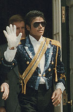 A mid-twenties African American man wearing a sequined military jacket and dark sunglasses. He is walking while waving his right hand, which is adorned with a white glove. His left hand is bare.