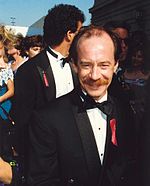 A smiling, dark-haired, balding man with a large mustache, wearing a tuxedo.  He is wearing a red AIDS ribbon on his left lapel.