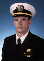 Color picture of Michael Murphy, a white male, wearing a military white dress hat and dark blue suit. There is a blue background behind him and he is wearing a gold Navy Seal Trident, two blue and green striped ribbons, one red and yellow striped ribbon and gold parachute insignia wings below the ribbons.